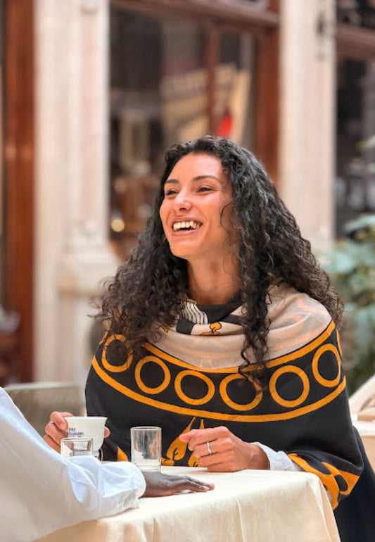 Woman in a patterned sweater sitting at a table, smiling outdoors.